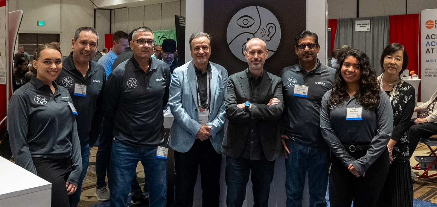 チームのメンバー8名がブースを背景にこちらに向い笑顔でこちらをみている。