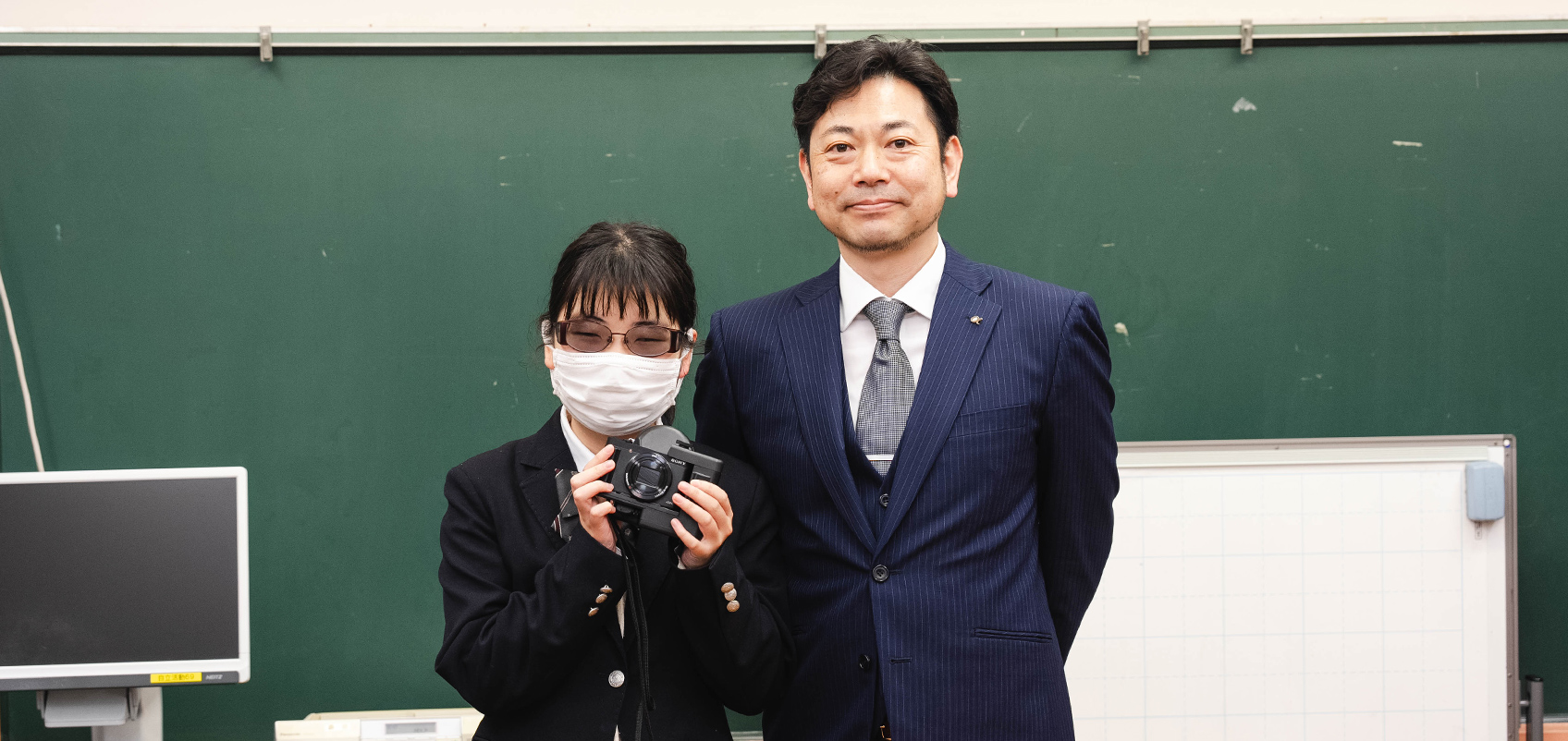 学校の教室で、黒板を背景に立つ、スーツにネクタイ姿の男性と制服の女子生徒の写真。女子生徒はマスクを着け、網膜投影カメラキットHX99 RNV を持っている