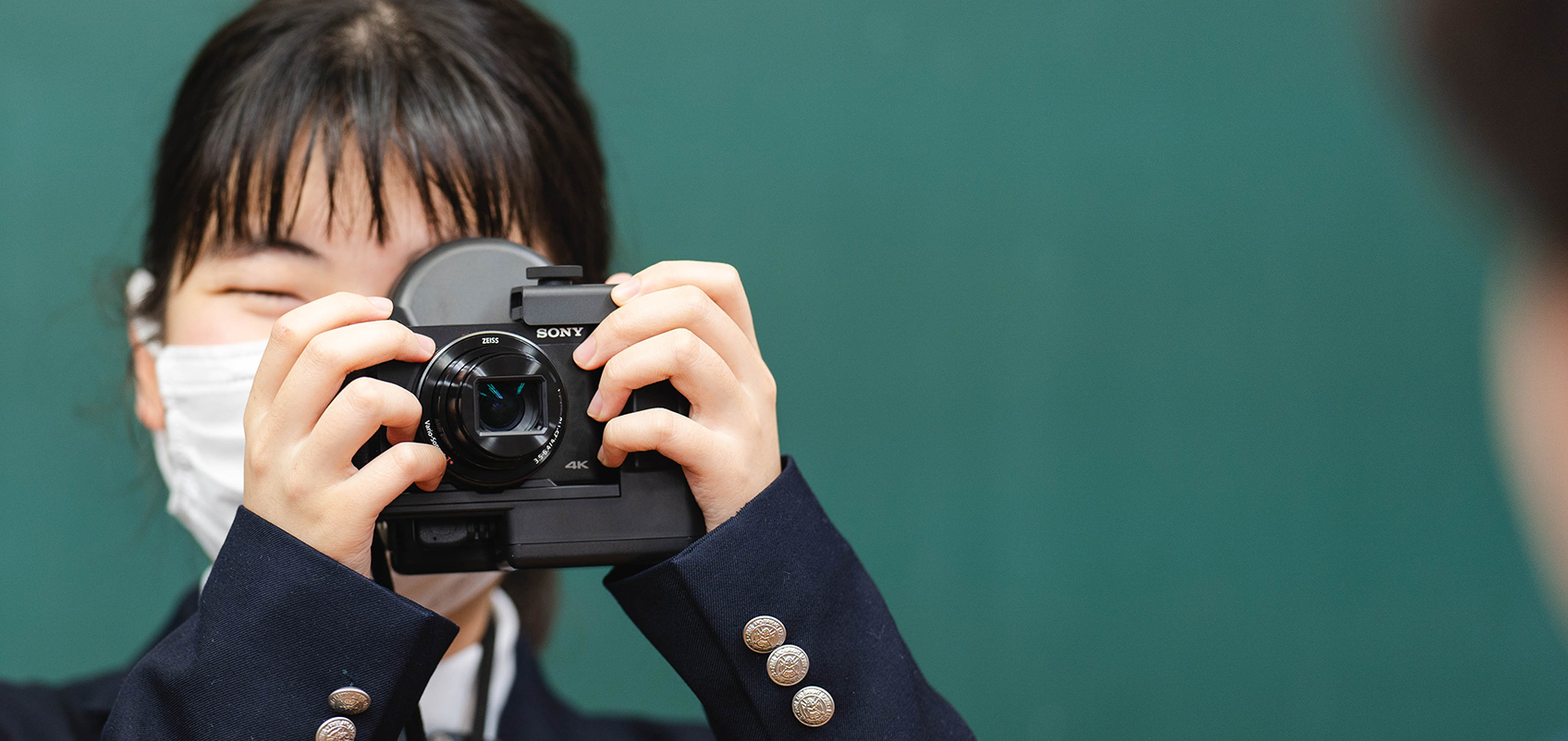 学校の教室で、黒板を背景に撮影された女子学生の写真。片目をつぶり、画面手前側の人物に向けて両手に持っている網膜投影カメラキットをのぞいている。