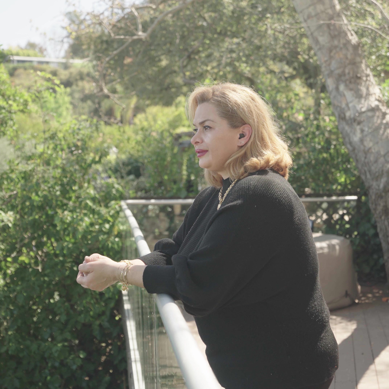 暖かい日差しの中、補聴器をつけた女性が木々に囲まれたバルコニーから景色をみている