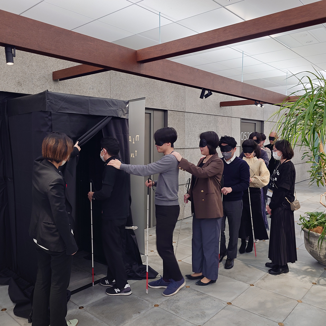 入口の様子を捉えた画像。入口に向かって列を作り並んでいる人々は黒いアイマスクをつけ、それぞれ前の人の肩に手を置いている。