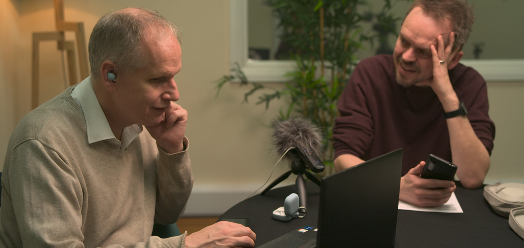 Image of two men sitting at a table with tripod-mounted microphone with furry wind screen between them. The man on the left has earbuds inserted and is using a PC, while the man on the right is watching him while holding a smartphone