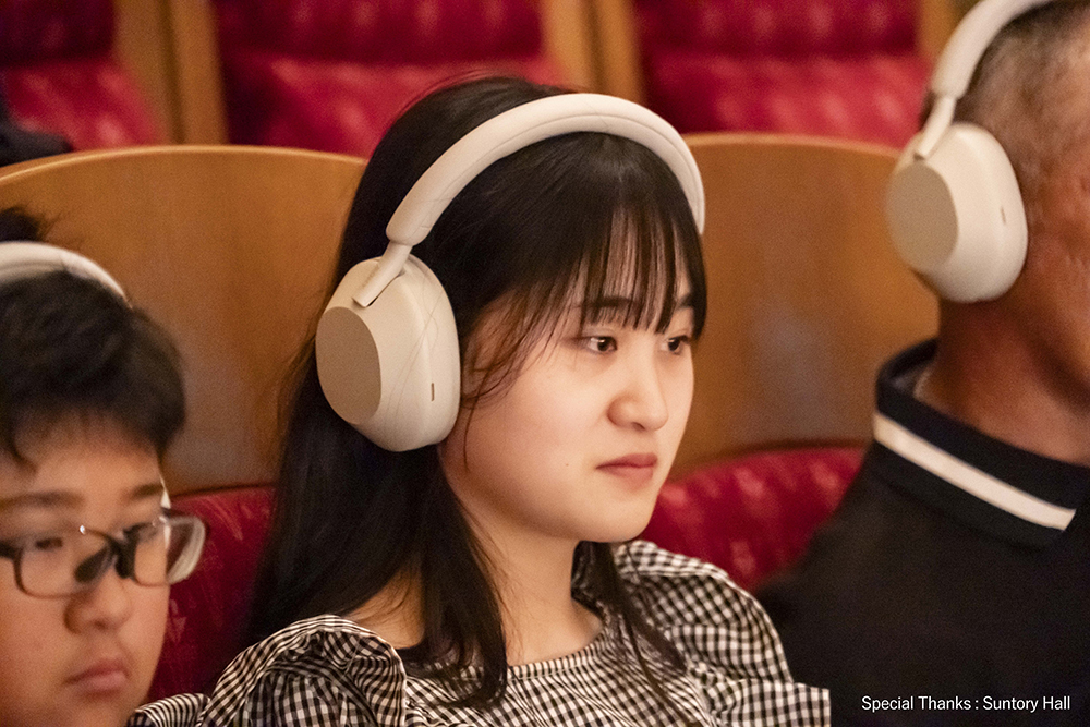 Three people sitting in a concert hall. In the middle is a girl, on the left side is a boy, and on the right side is a man. They are wearing white headphones. Special Thanks:Suntory Hall