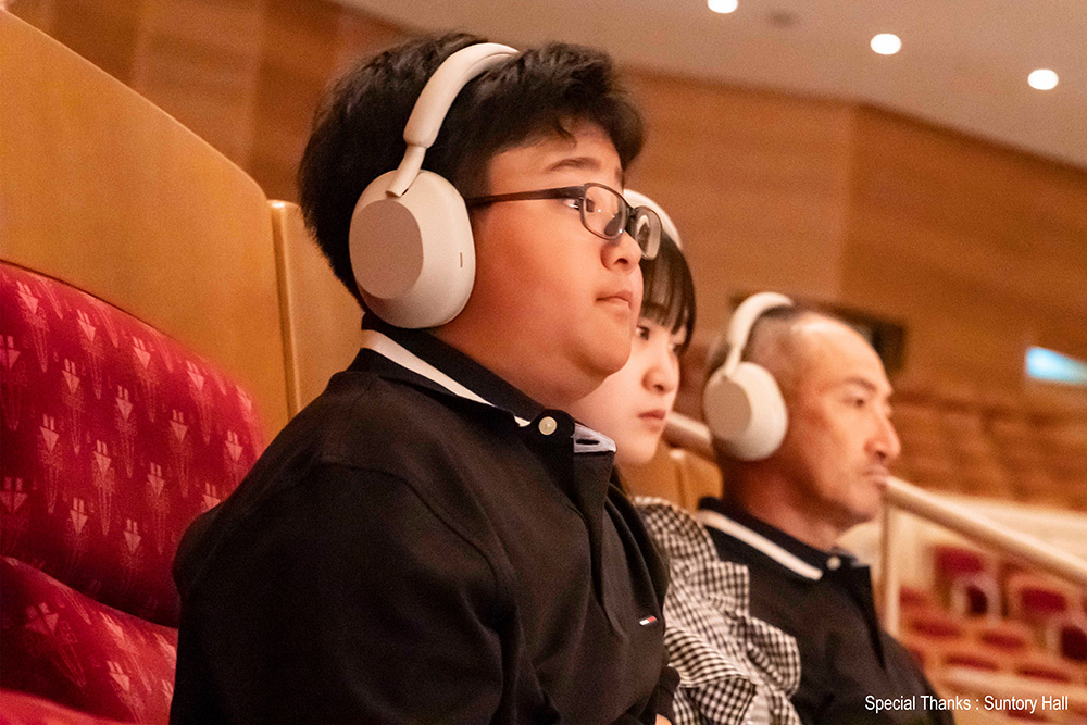 Three people sitting in a concert hall. From front to back, there is a boy, a girl, and a man. They are wearing white headphones. Special Thanks:Suntory Hall