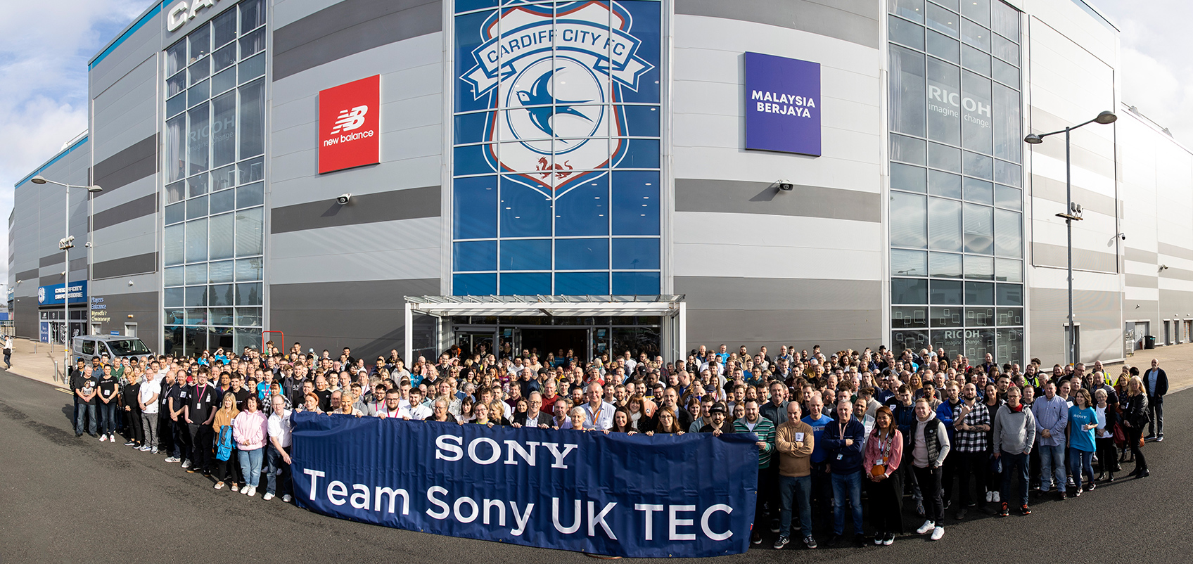 A large group of participants gathered in front of the building, holding a blue banner with the words "Team Sony UK TEC"